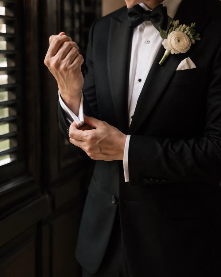 Groom in a tuxedo fastening his cuff beside a shuttered window